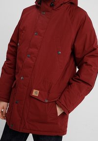 Rode geïsoleerde jas met een capuchon, voorzien van drukknoopsluiting, twee zijzakken en een merklabel aan de voorkant. Gladde stof met textuur.