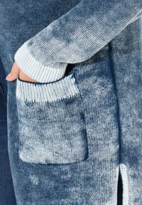 Strickjacke in einem Farbverlauf von Blau, mit langen Ärmeln, gerippten Bündchen und einer Vorderseite mit Tasche, weichem Material und geöffneten Seiten.