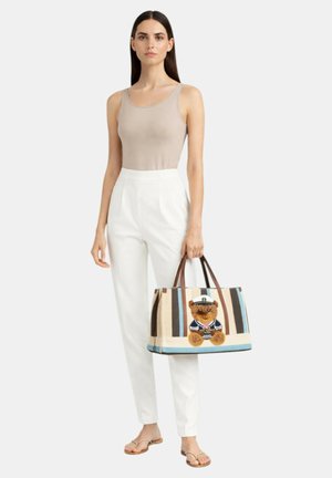 Kvinde i beige tanktop og hvide bukser, der holder en stribet taske med en teddybjørn iført sømandstøj.