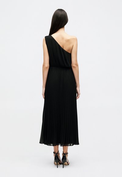 Γυναίκα που στέκεται με την πλάτη γυρισμένη, φοράει μαύρο πλισέ μίντι φόρεμα με έναν ώμο και μαύρα σανδάλια με ψηλό τακούνι, σε απλό φόντο.