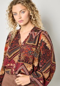 Langärmlige Bluse aus gemustertem roten, schwarzen und goldenen Stoff. Hat einen V-Ausschnitt, Knöpfe und eine fließende Passform. Mit gewelltem Randdetail.