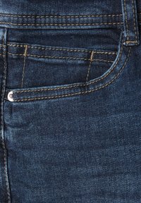 Tela de denim azul oscuro que muestra un bolsillo frontal con costura amarilla y un pequeño remache de metal en el lado izquierdo.