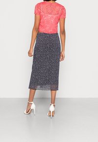 Haut rouge en dentelle à manches courtes associé à une jupe midi imprimée fleurs bleu marine. Le mannequin porte des sandales blanches à talons hauts, mettant en valeur une silhouette ajustée.