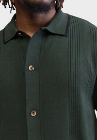 Gros plan sur un polo en maille vert foncé avec une texture côtelée verticale et deux boutons en écaille, porté par une personne aux dreadlocks courtes.