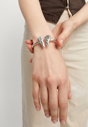 Brazalete de plata con acentos en forma de ala, con detalles texturizados, llevado en una muñeca. Banda lisa y pulida.