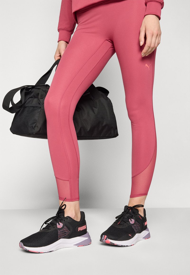 Schwarze Puma-Sportschuhe mit pinken Akzenten, kombiniert mit enganliegenden, rosafarbenen Leggings mit seitlichen Nähten und Netzdetails, gehalten mit einer schwarzen Sporttasche.