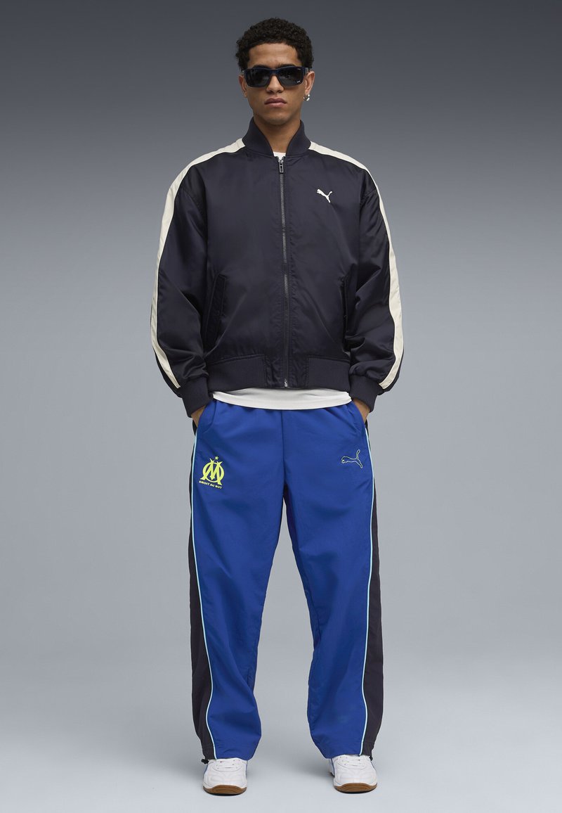 Puma bomberjakke i marineblå med cremefarvede striber, parret med blå sweatpants med neon Marseille-logo og sorte sidepaneler. Hvide sneakers.