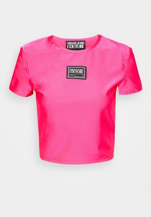 Różowa, satynowa, krótkoskrócona koszulka z krótkim rękawem, z czarną prostokątną metką marki pośrodku klatki piersiowej. Gładka faktura.