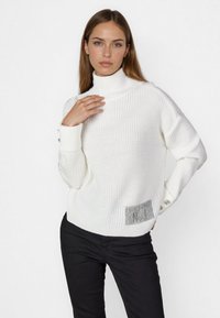 Weißer Strickpullover mit hohem Kragen, strukturiertem Stoff und einem silbernen Akzentpatch auf der unteren linken Seite. Slim Fit. Schwarze Hose.