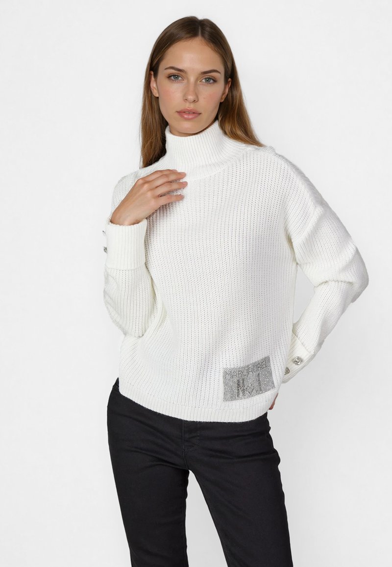 Weißer Strickpullover mit hohem Kragen, strukturiertem Stoff und einem silbernen Akzentpatch auf der unteren linken Seite. Slim Fit. Schwarze Hose.