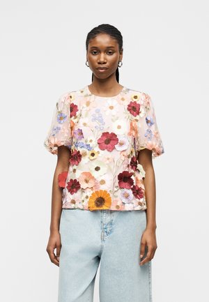 Femme portant une blouse transparente avec une broderie florale 3D colorée et des manches bouffantes, associée à un jean taille haute bleu clair.