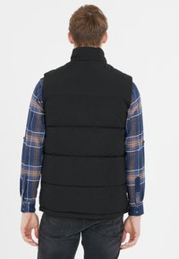Schwarze gepolsterte Weste mit hohem Kragen, zeigt gesteppten Abschnitte. Getragen über einem karierten blauen Hemd. Schlichte Rückseite ohne sichtbare Logos oder Beschläge.