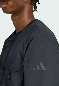 Detailný záber osoby v čiernej bunde s logom Adidas na hornej časti rukáva a s gombíkovou náprsní kapsou.