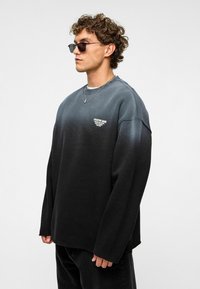 Zwarte gradient sweatshirt van zacht materiaal, met lange mouwen en een ronde hals. Bevat een klein logo op de borst.