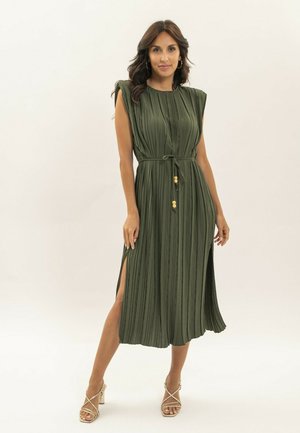 Une femme porte une robe midi sans manches vert olive avec des plis verticaux, une ceinture à la taille, des fentes latérales, et des talons beiges à brides.