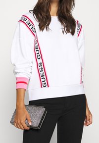 Sudadera blanca con la marca "GUESS" en rosa y negro en las mangas, puños acanalados y textura suave. Combinada con un bolso de mano negro con cuentas.