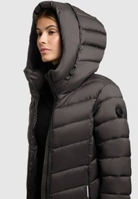 Bunda s kapucňou v tmavosivej farbe, s veľkou kapucňou, prešívaným vzorom, predným zipsom a logom na ľavom rukáve. Mäkkučký, izolačný materiál.
