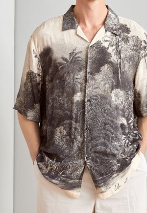 Homme portant une chemise à manches courtes beige avec un motif tropical en noir et gris complexe, assortie à un pantalon de couleur claire, les mains dans les poches.