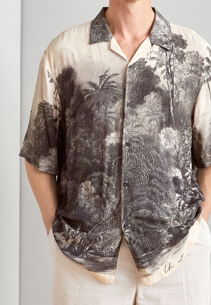 Uomo che indossa una camicia a maniche corte beige con bottoni, decorata con un intricato motivo tropicale in nero e grigio, abbinata a pantaloni chiari, con le mani nelle tasche.