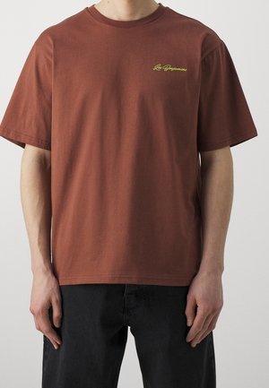 Person trägt ein schlichtes braunes T-Shirt mit kurzen Ärmeln und kleinem gelb-grünem Schriftzug "Los Benjamins" auf der oberen linken Brust.