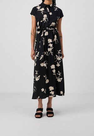 Femme portant une robe noire longueur cheville avec des motifs floraux blancs, un col, des manches courtes et une ceinture nouée à la taille, associée à des sandales noires.