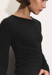 Profil egy fitted fekete, hosszú ujjú felsőről, oldalán redőkkel, világos háttér előtt.