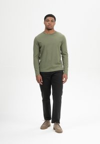 Langärmliges olivgrünes T-Shirt, schwarze Hose und beiges Schuhwerk. Das Shirt hat einen runden Ausschnitt und eine enge Passform, mit einer glatten Textur.