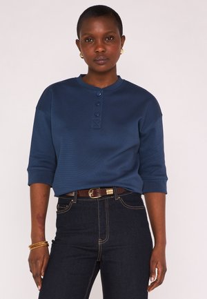 Donna con capelli corti che indossa una camicia blu navy con bottoni e jeans scuri con cintura marrone, in piedi davanti a uno sfondo chiaro uniforme.