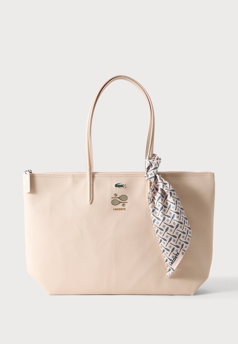 Sac cabas beige avec deux poignées, logo Lacoste, petits motifs sur le thème du tennis, et une écharpe blanche avec des motifs bleu Lacoste attachée à une poignée.