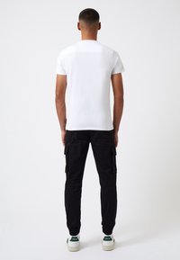 Camiseta de algodón blanca de manga corta, combinada con pantalones cargo negros con bolsillos laterales. Las zapatillas tienen acentos verdes y suelas blancas.