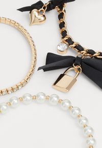 Three bracelets: one with gold chain and heart charm, one with faux pearls, and one metallic with crystals, featuring a lock and black ribbon.