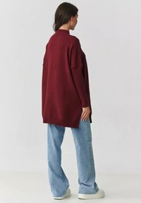 Burgundowy, oversize sweter z wysokim kołnierzem, prążkowanymi detalami i bocznymi rozcięciami, w połączeniu z jasnoniebieskimi spodniami typu wide-leg i białymi butami.