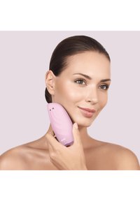 Dispositif de nettoyage facial rose avec une forme incurvée et une surface texturée, tenu contre la joue, présentant deux petits capteurs et un logo à la base.