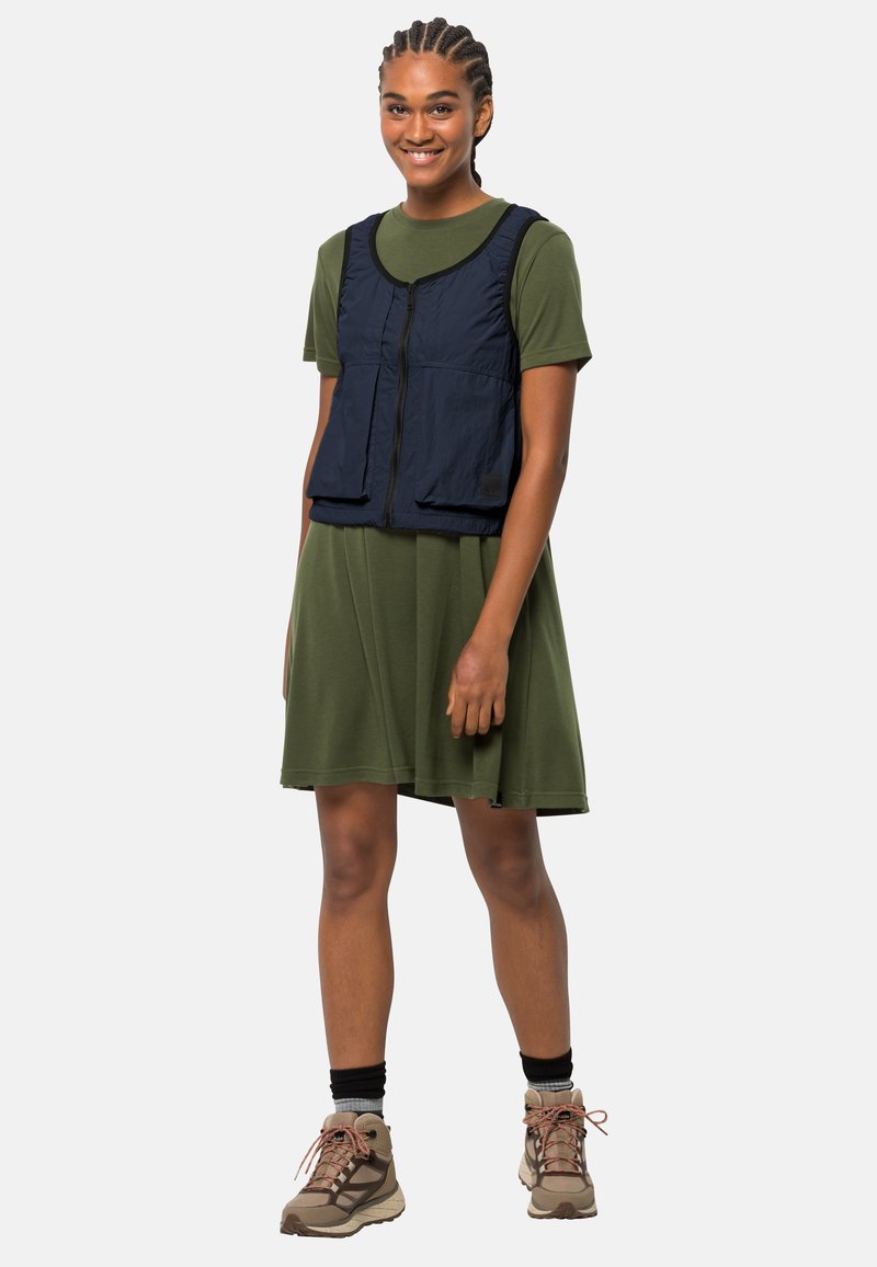 Granatowa bezrękawnik na zielonej sukience, z zamkiem błyskawicznym z przodu i dwoma kieszeniami. Model nosi szare buty turystyczne z czerwonymi sznurowadłami.