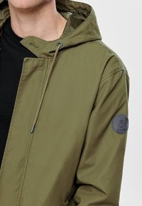 Olivgrön lättviktsjacka med huva, featuring ett logopatch på ärmen, tillverkad av ett slätt tyg med dragskojusteringar.