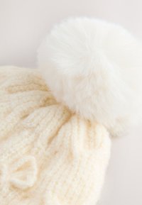 Cappello a cuffia in maglia di lana color crema con costine orizzontali e un grande pompon di pelliccia sintetica bianca e soffice sulla sommità. Texture morbida, design caldo.