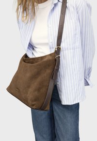 Hnědá semišová kabelka přes rameno s měkkou texturou, navržená se širokým otevřením a hnědým koženým páskem s kovovou přezkou.