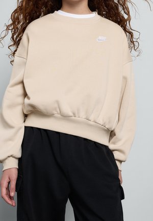 Person iført en beige Nike sweatshirt med et hvidt logo på brystet og løse sorte bukser foran en ensfarvet baggrund.