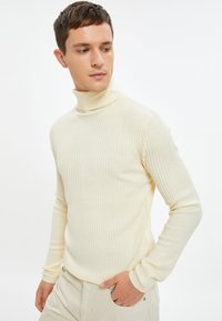 Cremfarbener, gerippten Rollkragenpullover mit langen Ärmeln, weicher Textur und taillierter Passform. Kombiniert mit hellen Hosen, die die Form des Oberteils betonen.