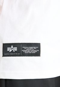 Valge T-särk musta ristküliku kujulise sildiga, kus on valge tekst ja logo, mis näitab brändi ja tootmisandmeid.