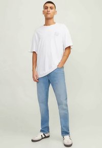 T-shirt de algodão branca com um corte relaxado e um pequeno gráfico no lado esquerdo do peito, combinada com jeans azul claro e ténis brancos com pormenores pretos.