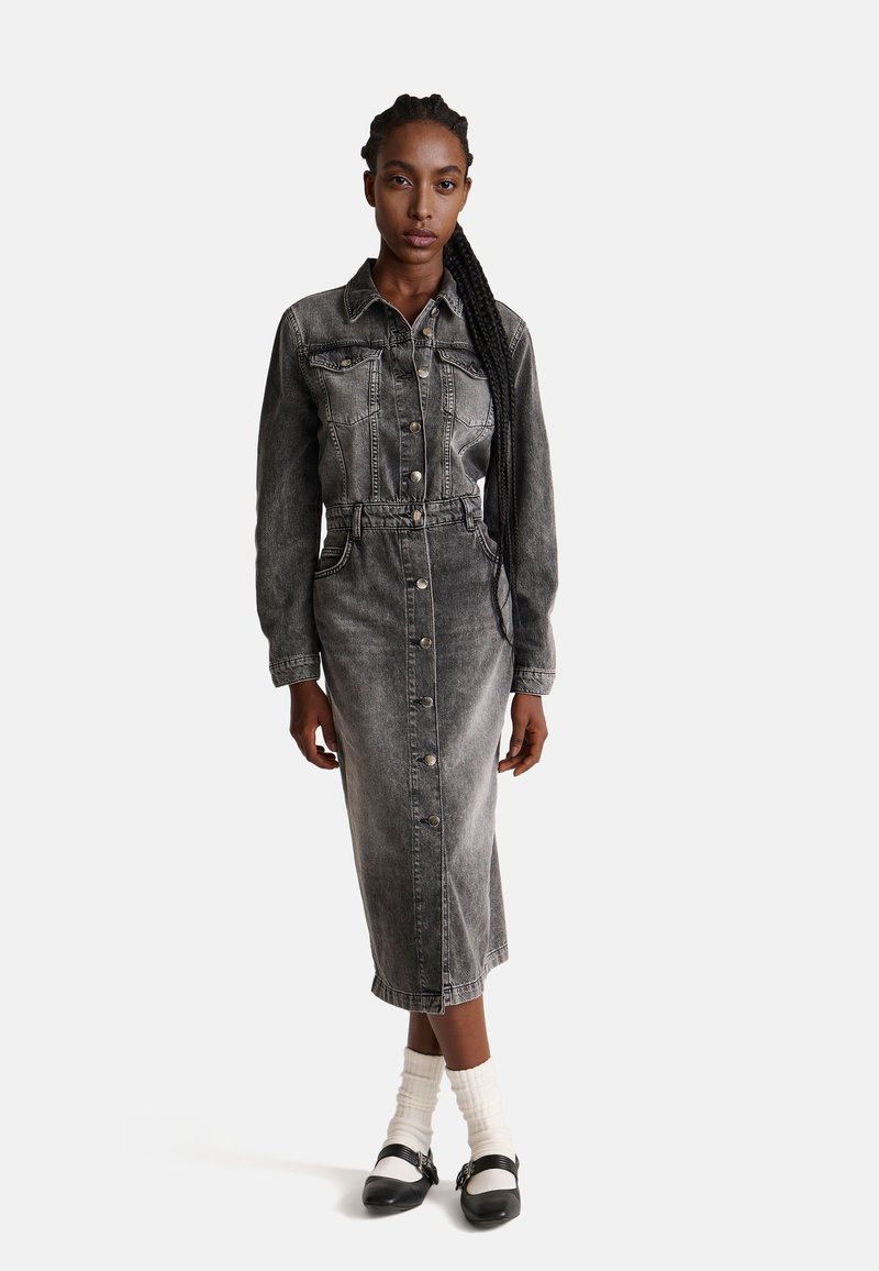 Szara sukienka dżinsowa z długimi rękawami, zapinana na guziki, z paskiem w talii. Posiada kieszenie na piersi i długość do połowy łydki. Noszona z białymi skarpetkami i czarnymi butami.