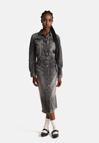 Abito in denim grigio con maniche lunghe, apertura con bottoni e vita con cintura. Presenta tasche sul petto e lunghezza fino al polpaccio. Indossato con calzini bianchi e scarpe nere.