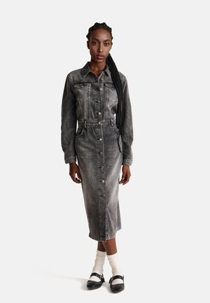 Grijze denim jurk met lange mouwen, knoopsluiting aan de voorkant en een ceintuur in de taille. Heeft borstzakken en een midi-lengte tot halverwege de kuit. G gedragen met witte sokken en zwarte schoenen.