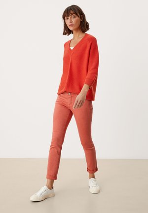 Pull en tricot orange vif avec un col en V et des manches 3/4, associé à un jean corail ajusté. Le modèle porte des baskets blanches.