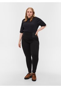 Haut noir à manches courtes associé à un pantalon noir taille haute avec des accents verts. Chaussures brunes à lacets avec une semelle épaisse.
