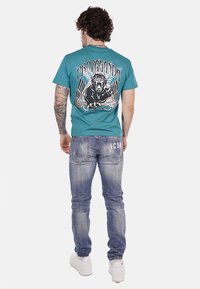 T-shirt in cotone verde acqua con stampa grafica di un leone sulla schiena, abbinato a jeans denim azzurri chiari con dettagli strappati e sneakers bianche.
