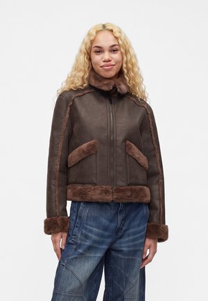 Jeune personne aux longs cheveux blonds bouclés, portant une veste en peau retournée marron et un jean bleu ample, debout devant un fond uni.