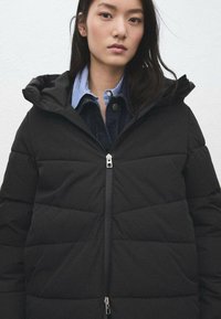 Czarna puchowa kurtka z poziomym pikowaniem, zapinana na zamek z przodu, z kapturem. Nosi się ją na niebieskiej koszuli w paski i dżinsach, charakteryzuje się smukłym krojem.