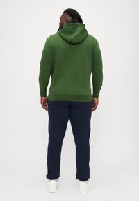 Zöld kapucnis pulóver sima textúrával, bordázott mandzsetta- és szegélyrészekkel, tengerészkék nadrággal párosítva. A lábbeli fehér sportcipő.